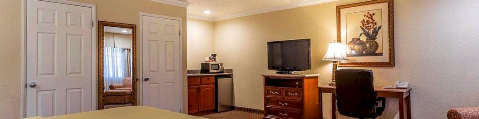 The image shows a hotel room with a queen-sized bed, TV, desk, chair, microwave, mini-fridge, and armchair with a painting on the wall.