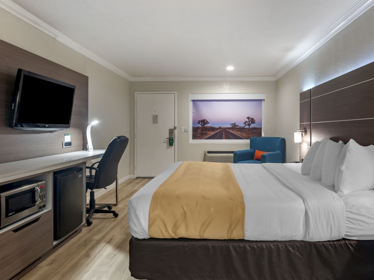 A modern hotel room features a bed, desk with chair, wall-mounted TV, mini-fridge, and a seating area with artwork on the wall.