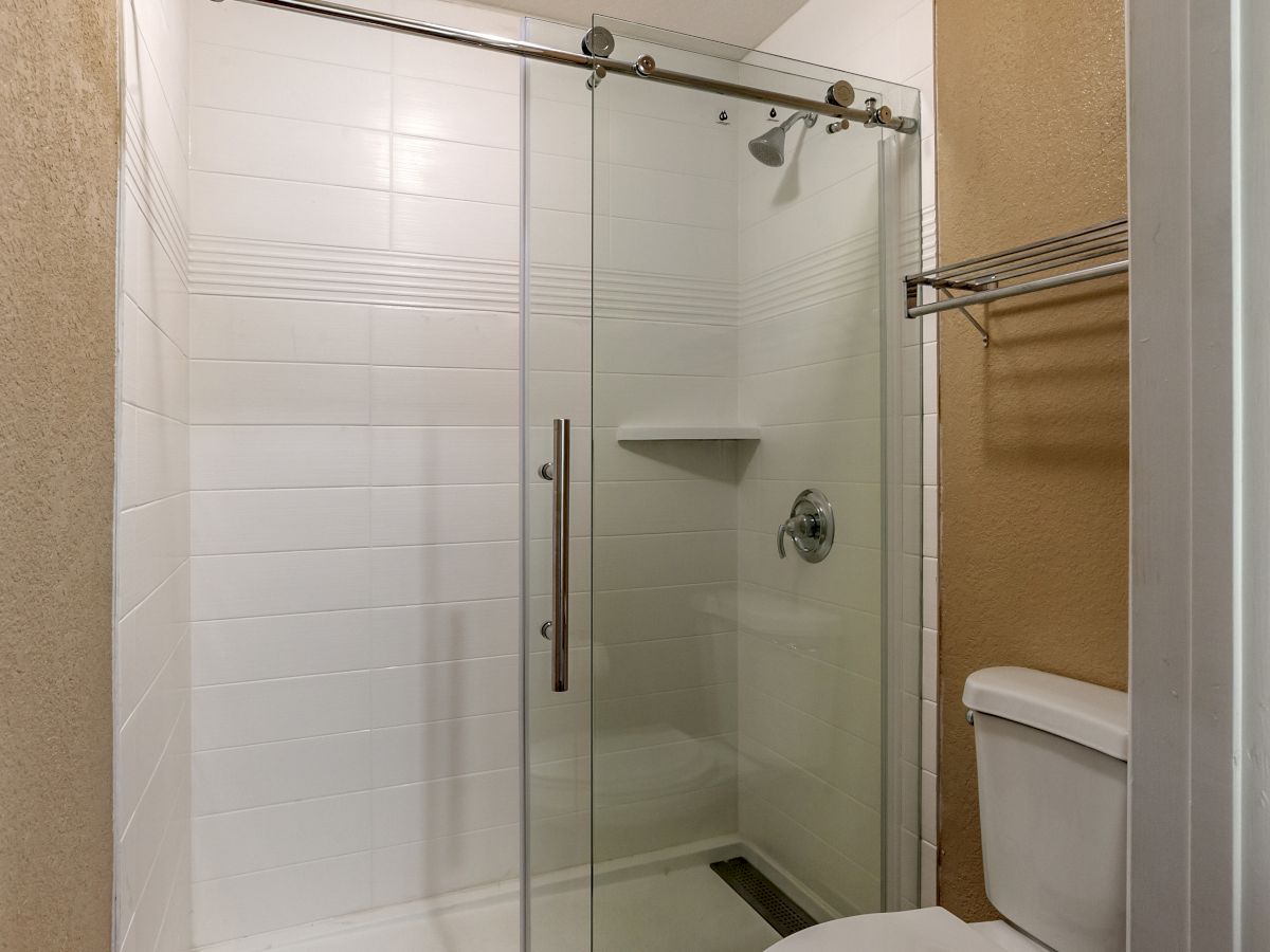 The image shows a bathroom with a glass shower enclosure, white tiled walls, a toilet, and a towel rack. The shower has a sliding glass door.