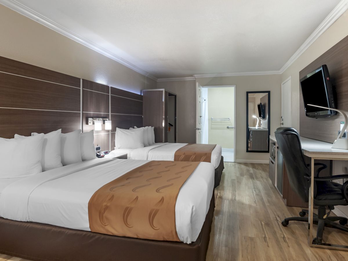 The image shows a hotel room with two double beds, a desk with a chair, a wall-mounted TV, and a bathroom in the background.