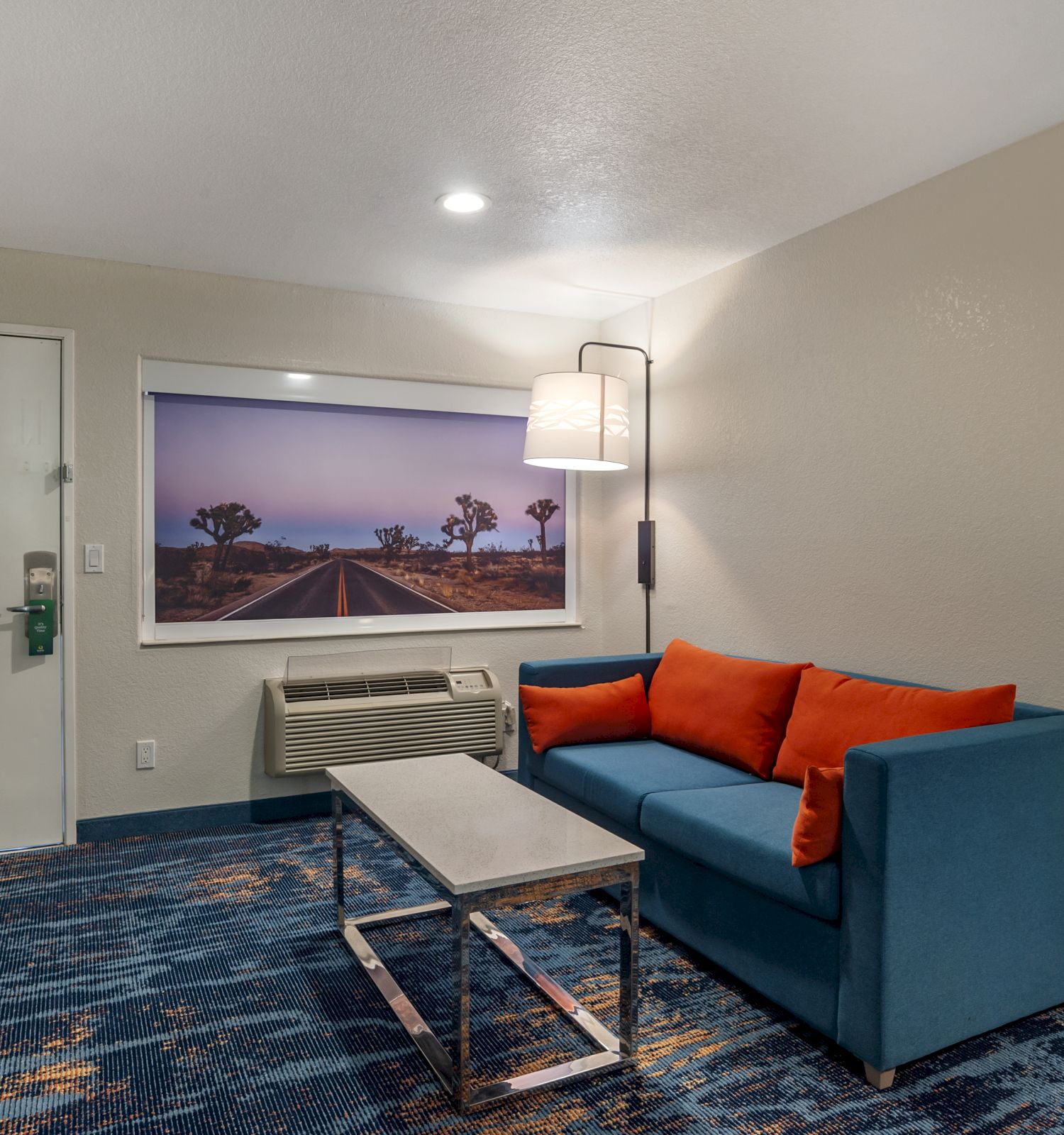 The image shows a cozy room with a blue sofa, orange cushions, a coffee table, a wall lamp, and a landscape photo. The room has a modern style.