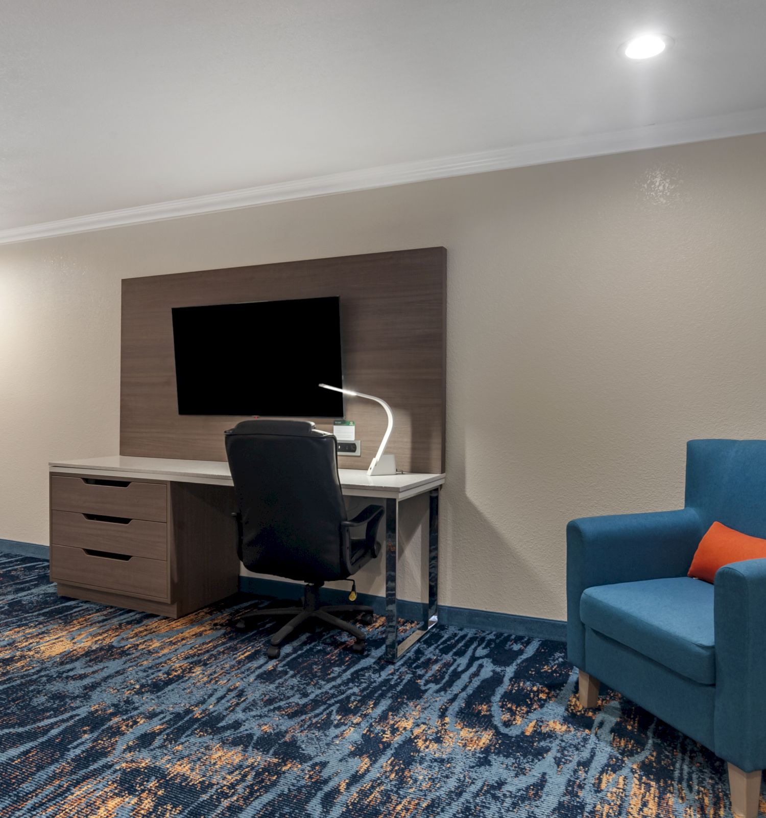 The image shows a modern hotel room with a desk, chair, TV, blue armchair, microwave, and a window with a view.