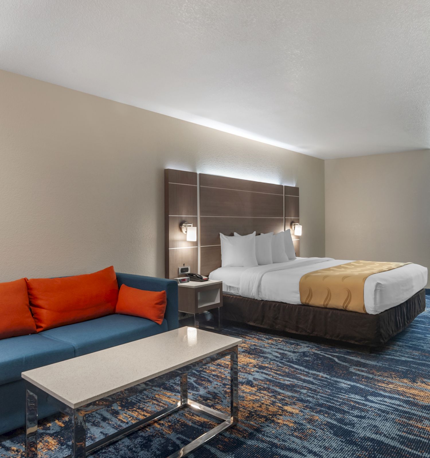 A hotel room with a bed, blue sofa with orange cushions, a coffee table, a desk and chair, and a closet area in the background.