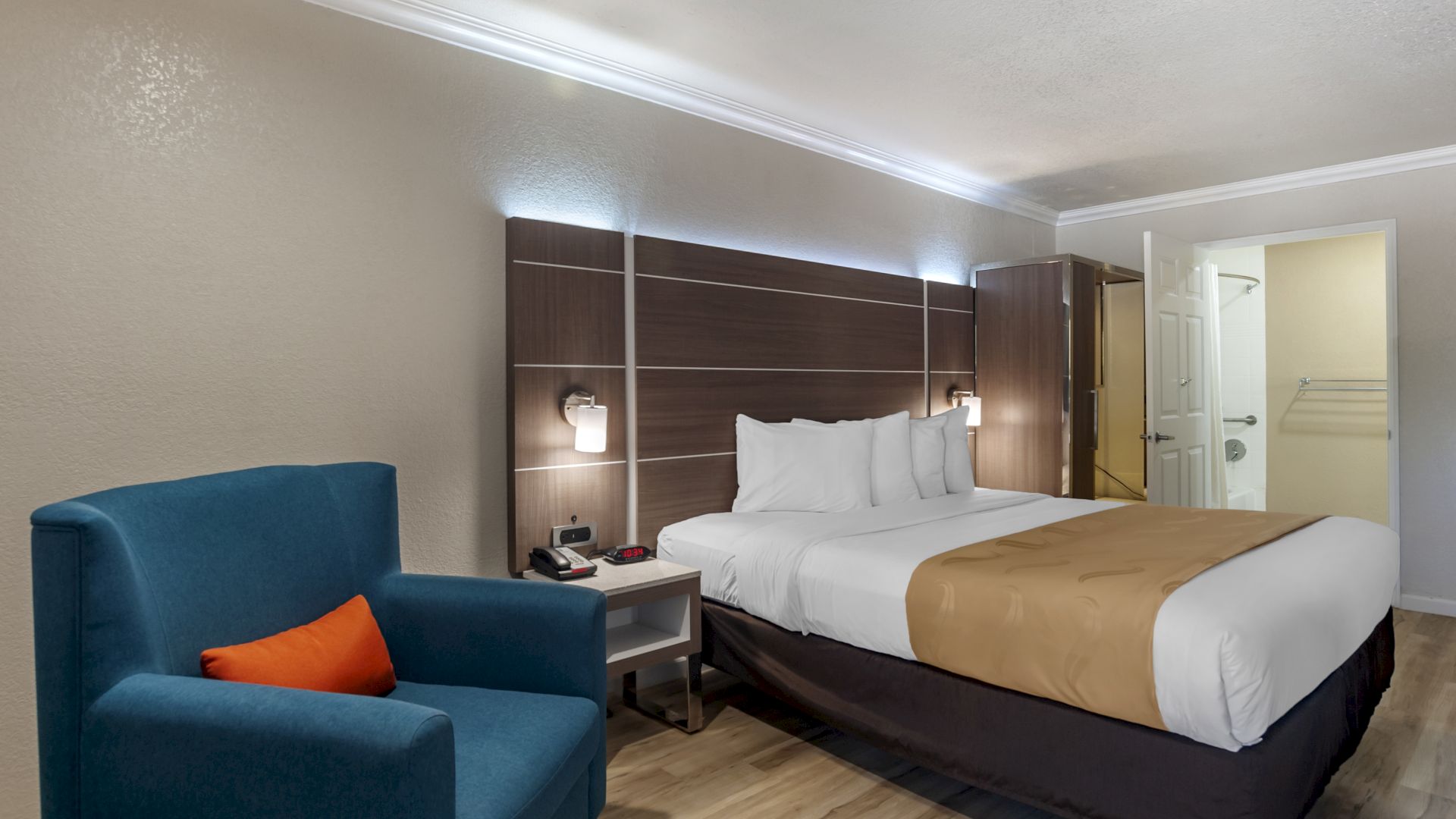 A neatly made bed in a modern hotel room with a blue armchair, wooden flooring, and wall-mounted lights, with an open door to the bathroom is visible.