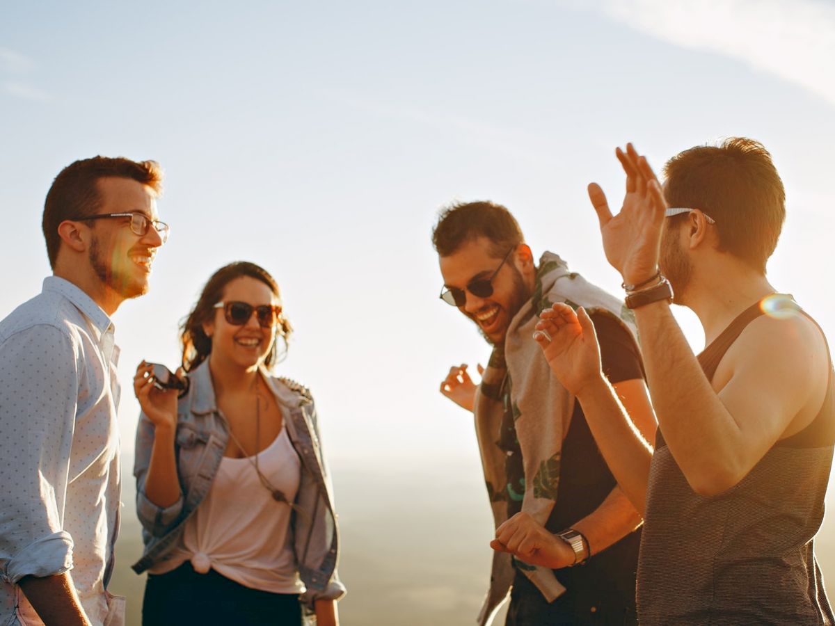 A group of four people is outdoors, smiling and enjoying each other’s company in the sunlight, some wearing sunglasses.