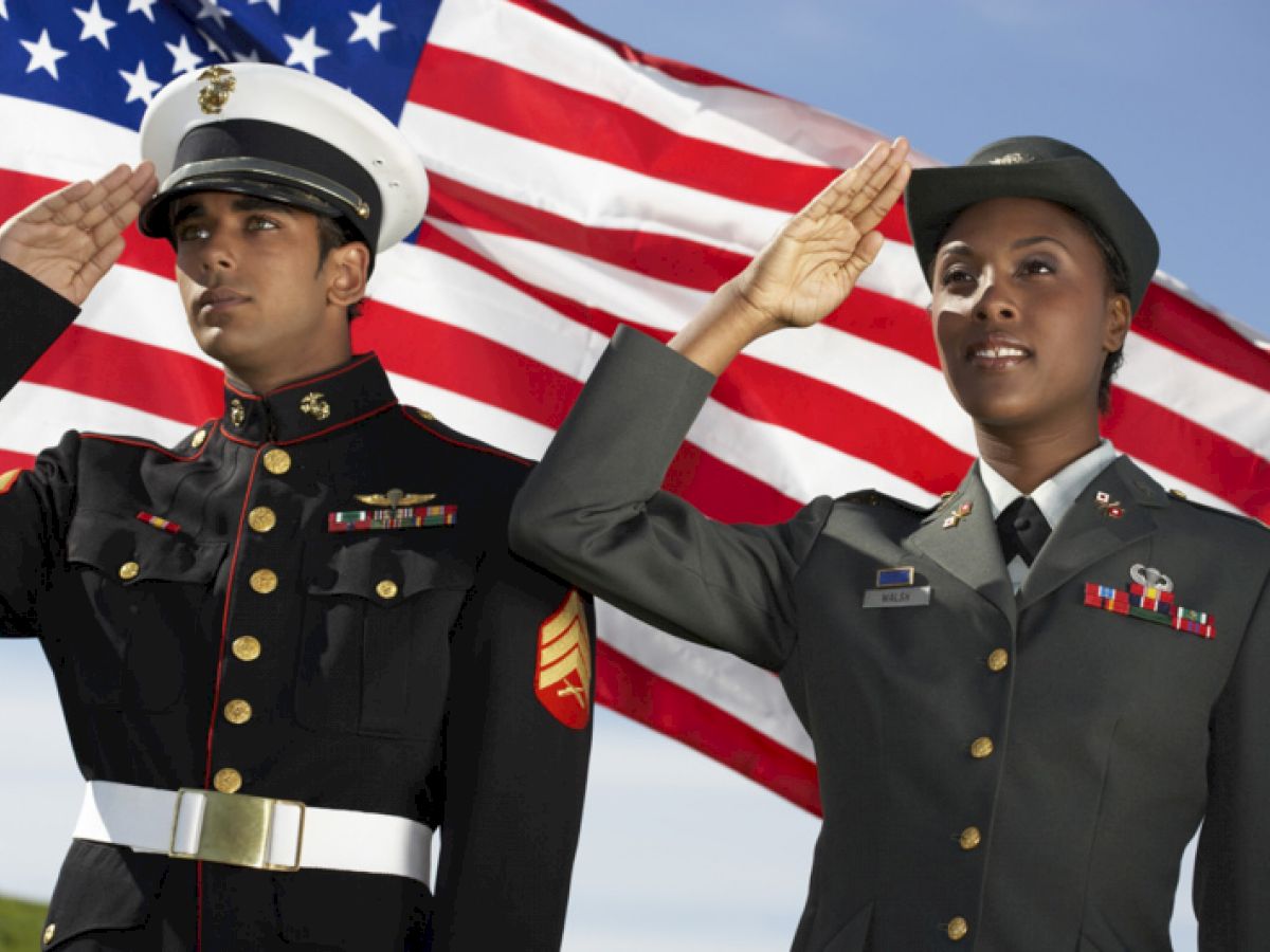 Two uniformed military personnel saluting in front of a waving American flag.