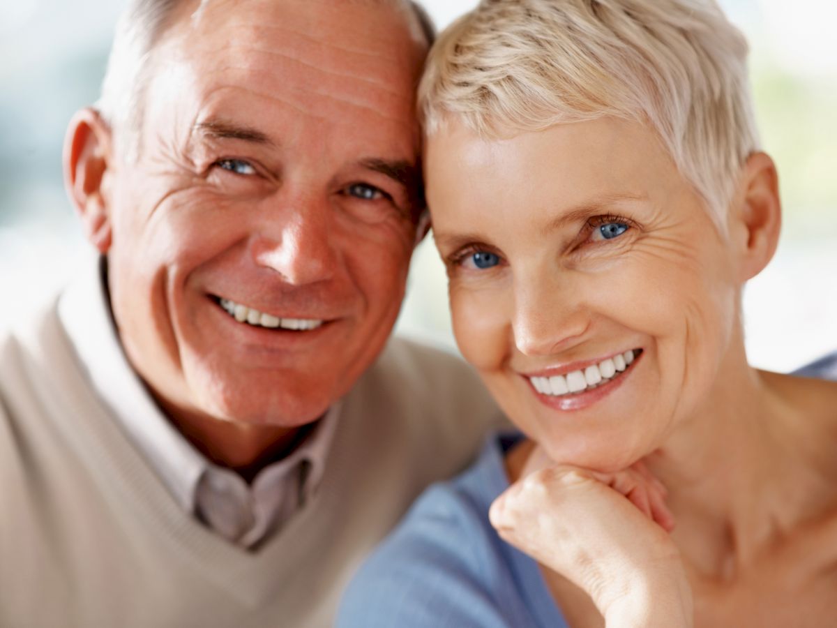 The image shows a smiling older couple, with the man wearing a beige sweater and the woman with short blonde hair, posing closely together.