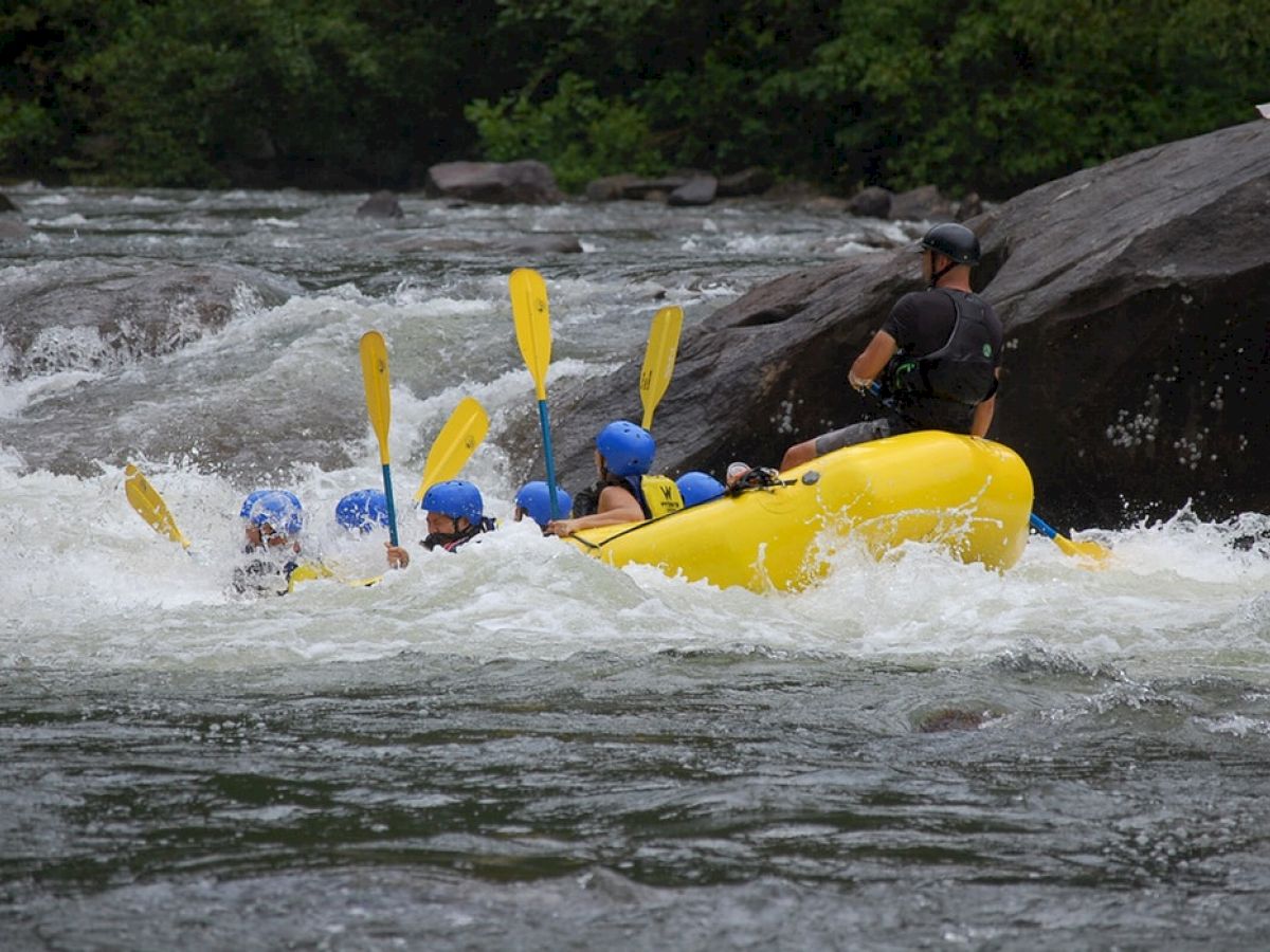 A group of people in blue helmets are white-water rafting in a yellow raft, paddles raised, navigating through rough waters and rocks.