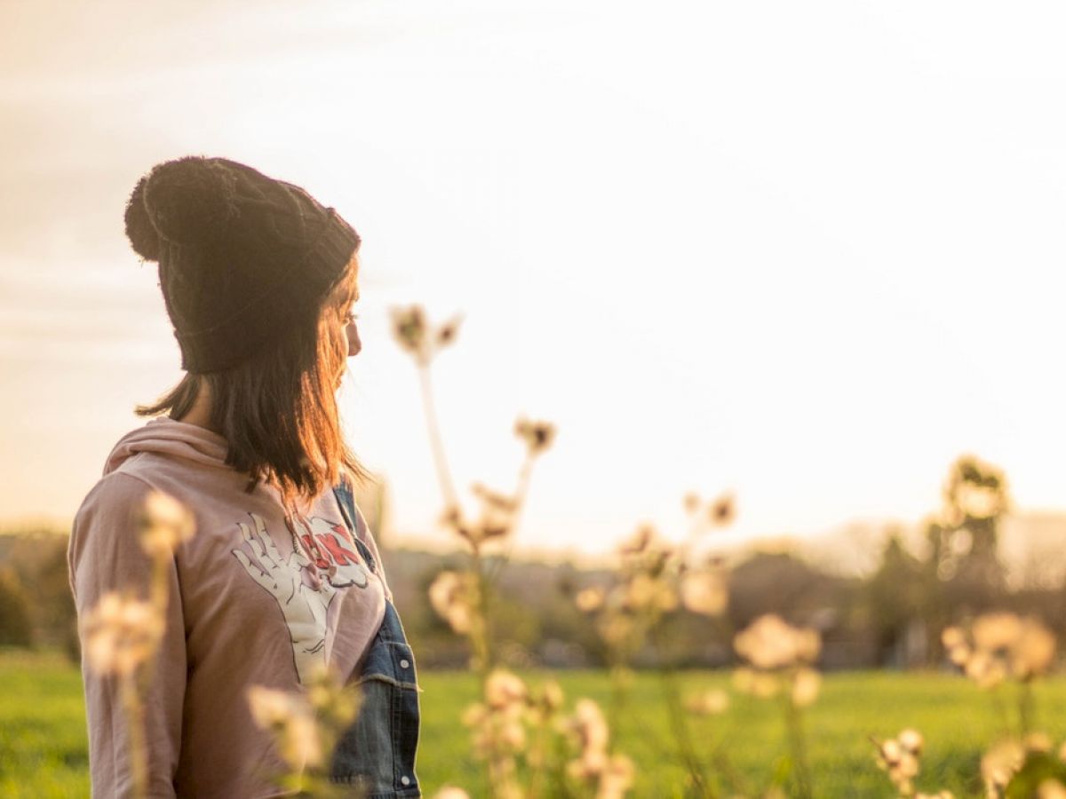 A person wearing a beanie and jacket stands in a field of flowers, looking towards the sunset with a bright sky in the background.