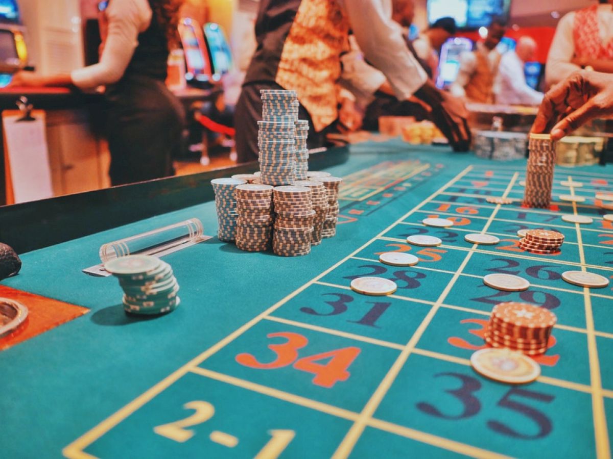 A casino roulette table with various chips stacked and scattered, and casino staff assisting players in the background.