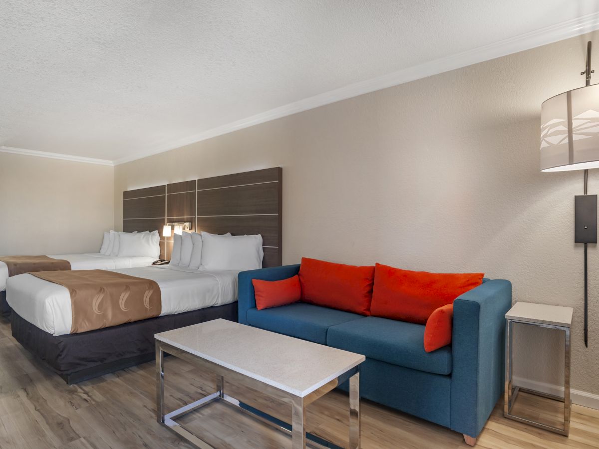 A hotel room with two beds, a blue sofa with red cushions, a coffee table, and modern lighting on a wooden floor.