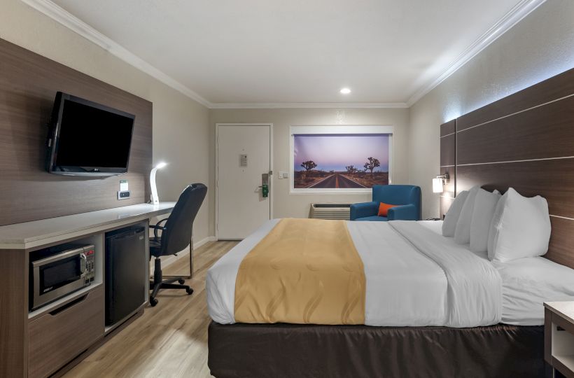 The image shows a neatly arranged hotel room with a bed, a wall-mounted TV, a desk with a chair, a microwave, a mini-fridge, and a picture of a road.