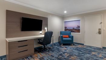 The image shows a hotel room with a wall-mounted TV, a desk and chair, a blue armchair, and a window with a view of a sunset.