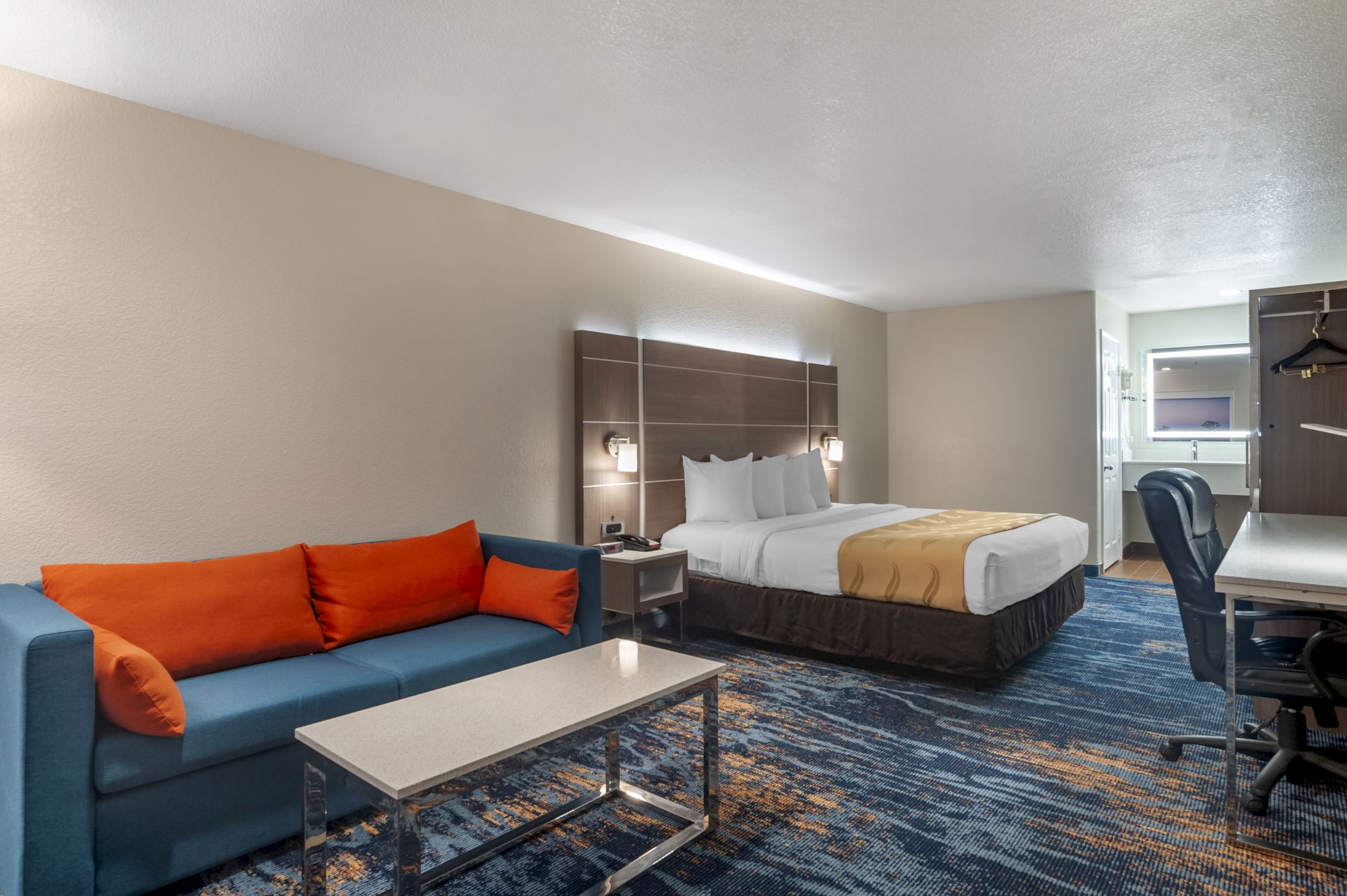 A hotel room features a bed with white linens, a blue sofa with orange pillows, a desk with a chair, and a coffee table on a patterned carpet.