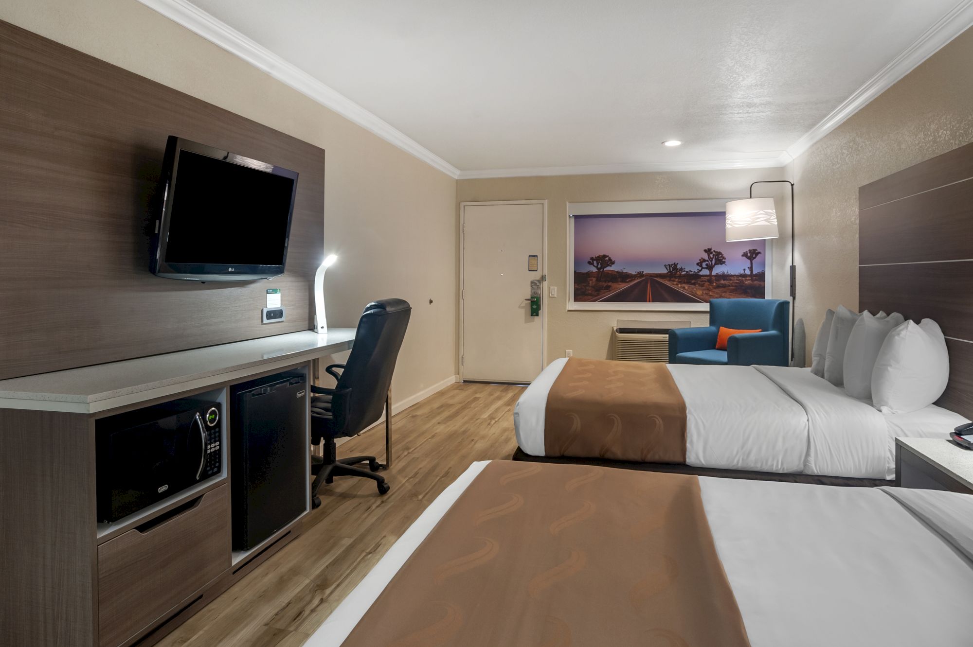 A hotel room with two beds, a wall-mounted TV, a desk with a chair, a mini-fridge, and a scenic picture above the air conditioning unit, ending the sentence.