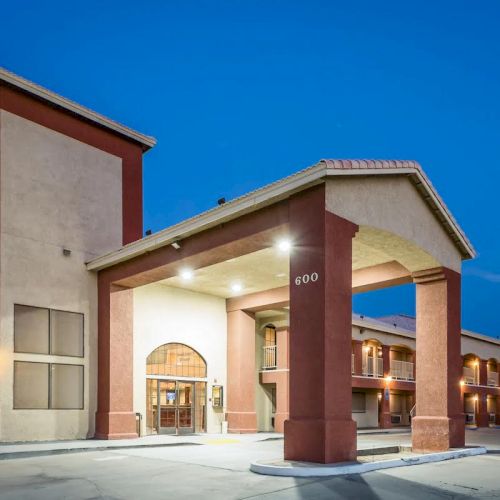 A modern hotel entrance with a tall porte-coch&egrave;re, warm lights, and a row of rooms extending into the distance at dusk.