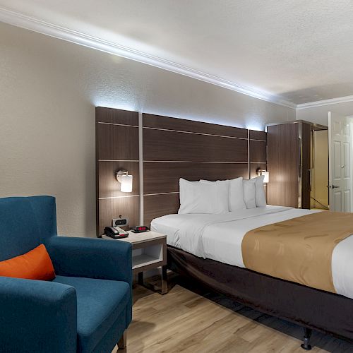 A modern hotel room with a large bed, blue armchair, bedside tables, lamps, and an open door leading to a bathroom.