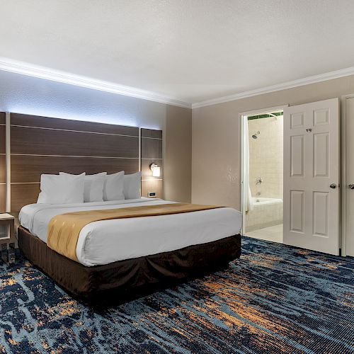 A modern hotel room with a large bed, bedside tables, lamps, and a blue-patterned carpet. An open door reveals a bathroom with a bathtub.