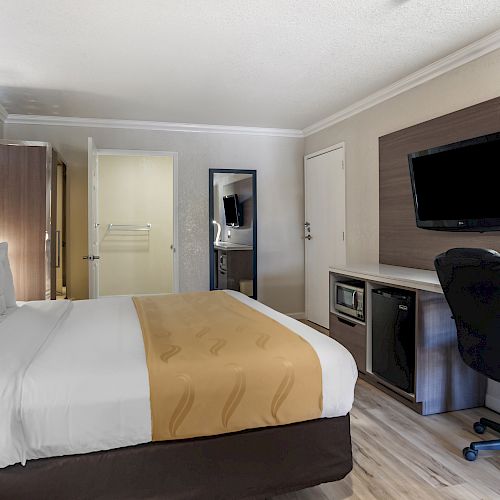 A modern hotel room with a queen bed, desk and chair, TV, mini-fridge, microwave, and a mirror; an open door reveals a bathroom.