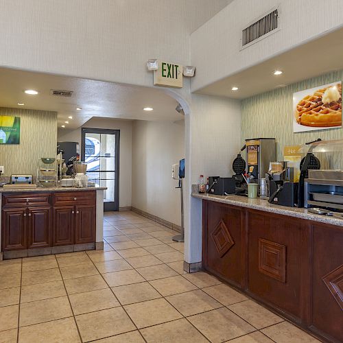 The image shows a small breakfast buffet area with food and drink dispensers, counters, and an exit sign above the doorway at the back.