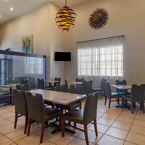 This image shows a modern dining area with tables, chairs, pendant lights, a wall-mounted TV, and a decorative sun-like wall piece.