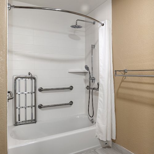 This image shows a bathroom with a white bathtub, chrome grab bars, an adjustable shower head, a shower curtain, and a towel rack on a beige wall.