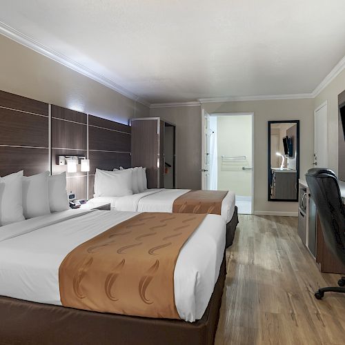 A clean, modern hotel room with two double beds, a TV, desk, chair, a closet, and a mirror, featuring wood flooring and minimalist decor.