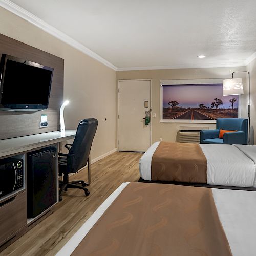 A hotel room with two beds, a wall-mounted TV, a desk with a chair, a mini fridge, and a large picture of a beach scene on the wall.