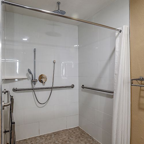 A spacious, accessible shower area with grab bars, a handheld showerhead, and a white curtain on the right side.