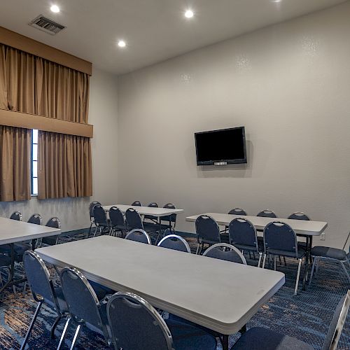 This image shows a small conference room with tables, chairs, a wall-mounted TV, a whiteboard on a stand, and a window with brown curtains.