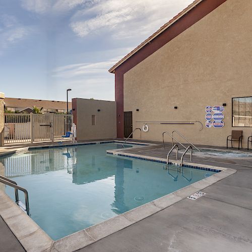 The image shows an outdoor swimming pool next to a building, with safety equipment, pool rules signs, and lounge chairs by the poolside.