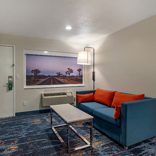 This image shows a hotel room with a blue sofa, orange cushions, a coffee table, wall art, a lamp, and a door.