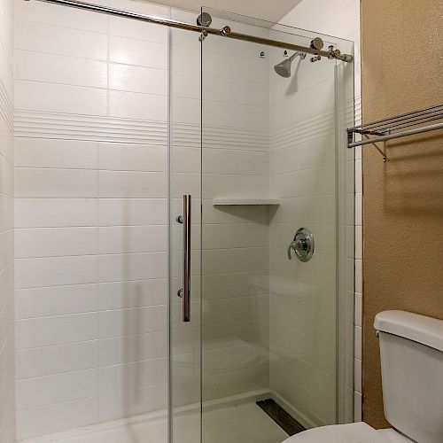 The image shows a bathroom with a glass-enclosed shower, white tiles, a chrome showerhead, a small shelf, and a toilet on the right.