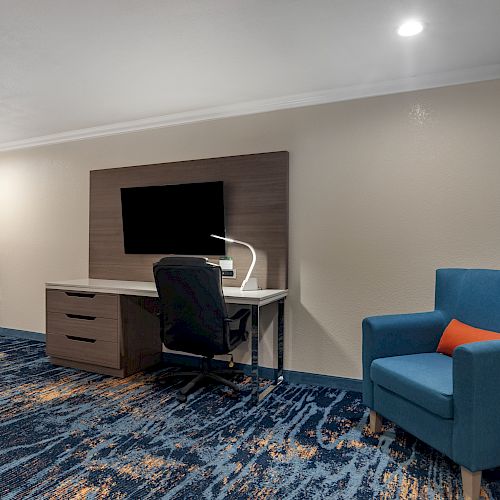 This image shows a modern room with a desk, chair, TV, lamp, armchair, and a small kitchenette with a microwave and fridge in the corner.