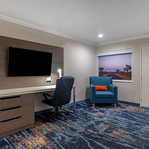 The image shows a hotel room with a wall-mounted TV, desk, chair, single armchair, and a window with a landscape. The room has modern decor.