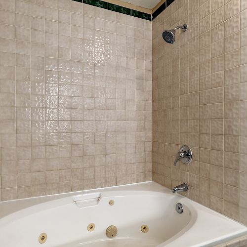 The image shows a bathroom corner with a beige tiled shower area and a white jacuzzi tub with jets. It features a showerhead and two faucets.