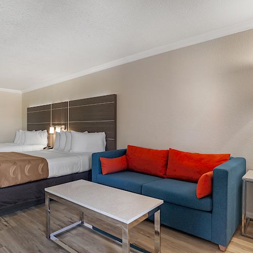 A hotel room with two double beds, a blue sofa with red cushions, a coffee table, and a wall-mounted lamp beside the sofa.