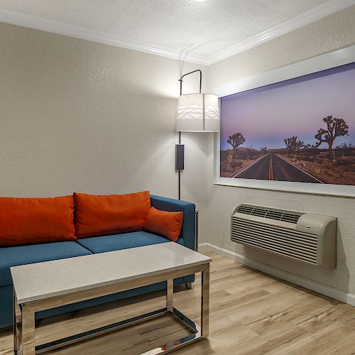 A cozy room with a blue couch featuring orange cushions, a wall-mounted lamp, a landscape picture, and a wall-mounted air conditioner, ending sentence.