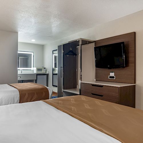 A hotel room with two beds, a wall-mounted TV, open wardrobe, and a sink area with a mirror in the background.