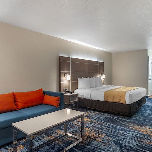 A hotel room with a bed, a blue sofa with orange pillows, a desk with a chair, and a table on a patterned carpet.