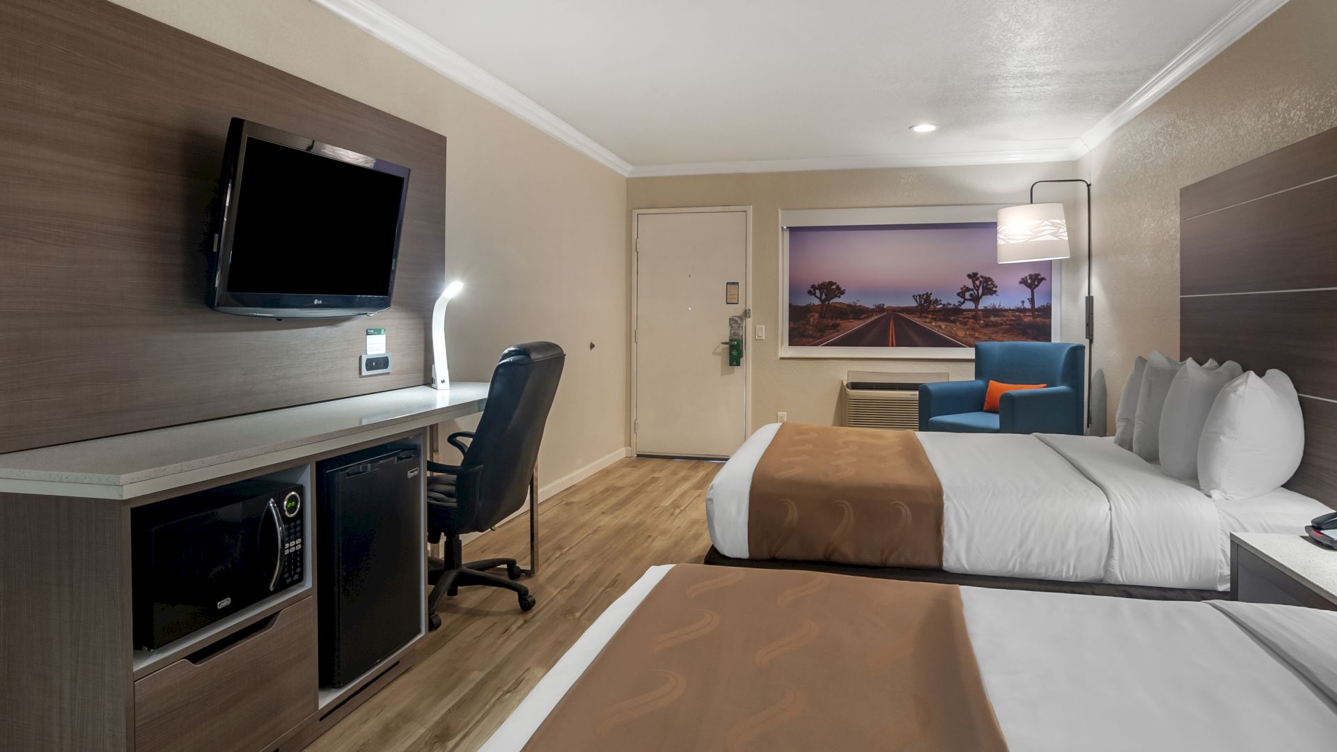 A hotel room with two beds, a TV, a desk with a chair, a mini-fridge, wood flooring, and a picture of a beach scene on the wall.