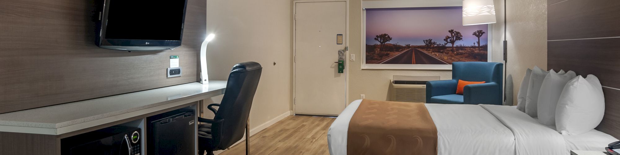 A hotel room with two beds, a TV, a desk with a chair, a mini-fridge, wood flooring, and a picture of a beach scene on the wall.