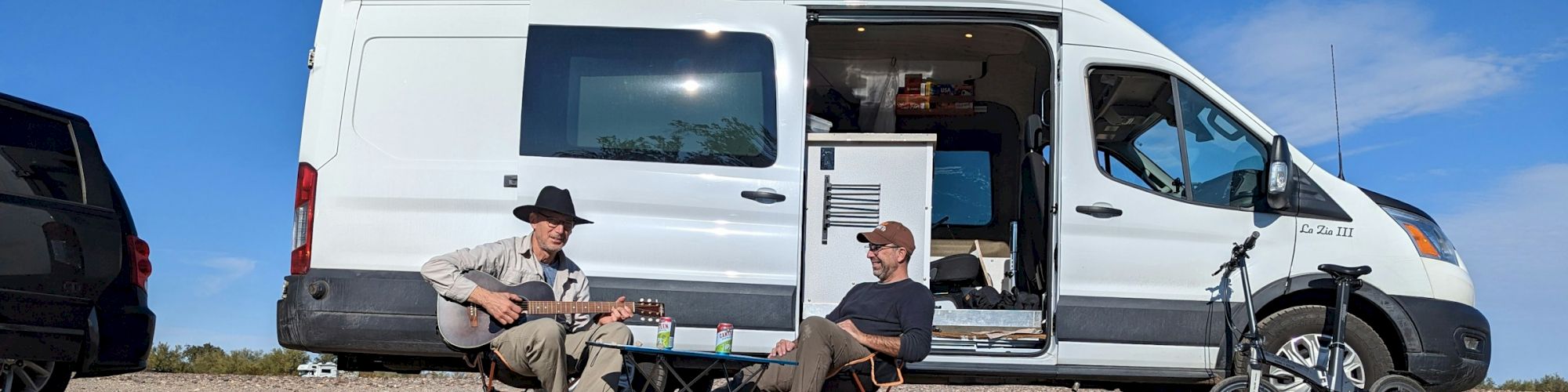 Two people are sitting outside a white camper van; one is playing guitar, while the other relaxes. A scooter is parked nearby, and it's sunny.