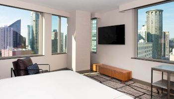 A modern hotel room with large windows showcasing a city skyline, a bed, a TV mounted on the wall, and minimalistic furnishings.