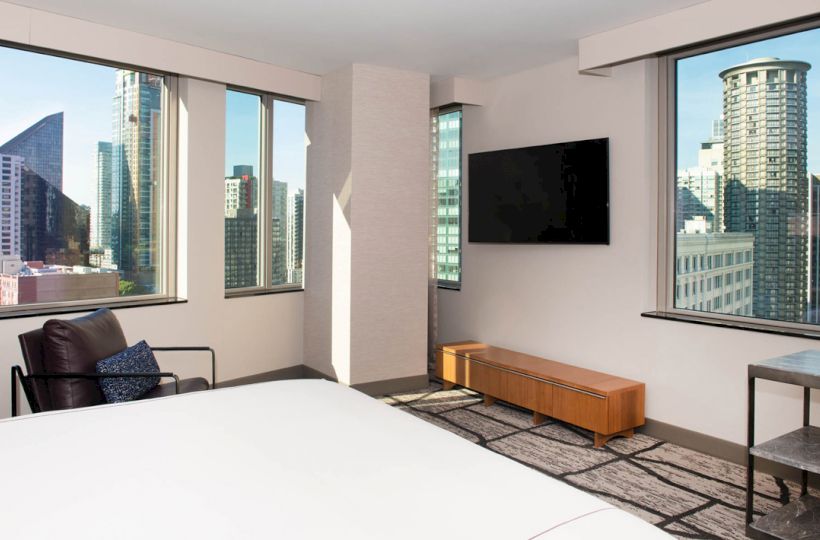 A modern hotel room with large windows showcasing a city skyline, a bed, a TV mounted on the wall, and minimalistic furnishings.