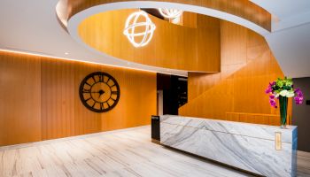 A modern lobby with a large wall clock, a marble reception desk, a curved wooden staircase, and decorative lighting and flowers.