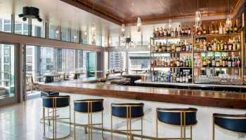 A modern bar with a wooden ceiling, hanging lights, well-stocked shelves of liquor, and high stools along a sleek counter, located in a city setting.