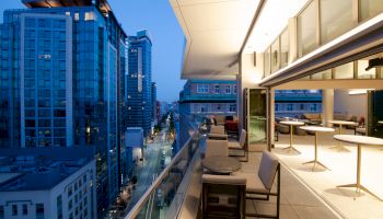 A modern, city apartment balcony with seating, overlooking tall buildings and a street in an urban setting at dusk.