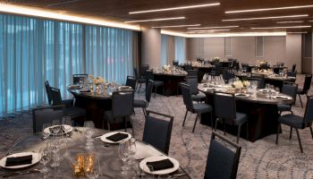 The image shows a modern banquet hall with several round tables set elegantly with plates, glasses, and floral centerpieces, ready for an event.