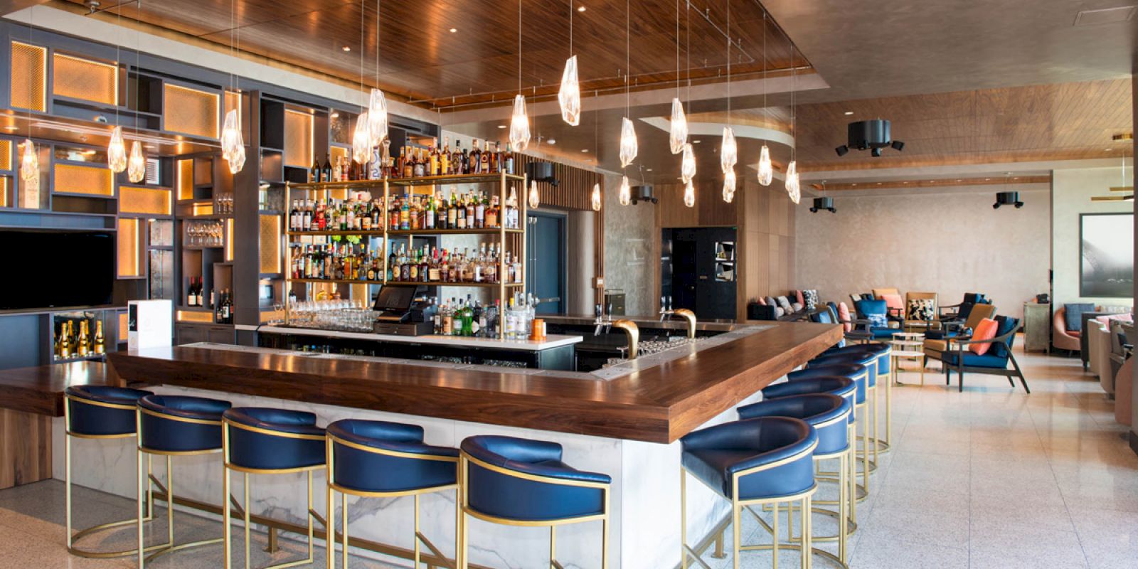 A modern bar with a wooden counter, blue bar stools, hanging lights, and a well-stocked shelf of bottles. Lounge area with seating in the background.